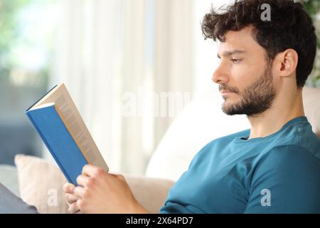Side view portrait of a serious man reading a paper book sitting on a couch at home Stock Photo