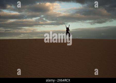 A woman is standing on a sandy beach, with the sky in the background. She is wearing a black dress and she is dancing. Scene is joyful and carefree Stock Photo