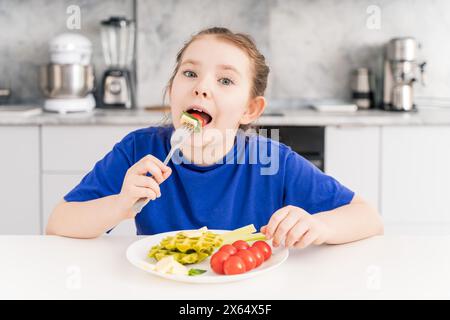 Happy little girl holding a fork in her mouth. A pre-teenage girl has breakfast in the kitchen at home with a plate of waffles, vegetables and cheese. Stock Photo