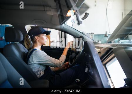 Female auto mechanic using diagnostic tool, scanner, running diagnostic on car. Beautiful woman sitting inside of car, performing diagnostic test. Stock Photo