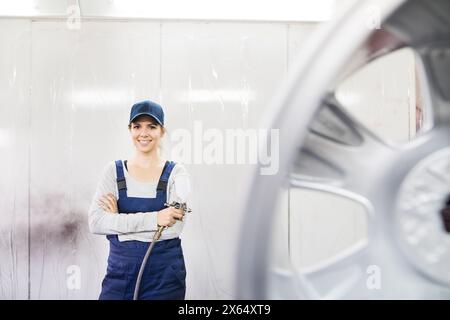 Female car painter, paingting vehicles in auto body shop. Young woman holding spray gun, spraying paint on rim of wheel, wearing coveralls. Stock Photo