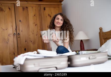 Beautiful woman planning summer vacation abroad, sitting on bed, packing suitcase. Solo female traveler looking at itinerary in tablet. Stock Photo