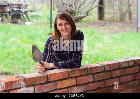 a young female bricklayer with a trowel in her hands is smiling, leaning against a brick wall Stock Photo