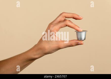 Close-up of a human hand elegantly presenting a coffee capsule, isolated on a beige backdrop Stock Photo