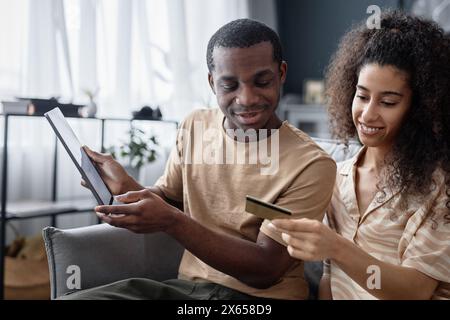 Medium shot of modern young biracial couple spending time at home doing online shopping in app on digital tablet Stock Photo