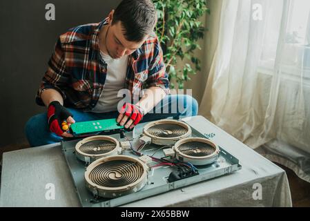 Concentrated technician in plaid shirt and red gloves repairs electric stove, carefully examining circuit board over disassembled appliance. Stock Photo