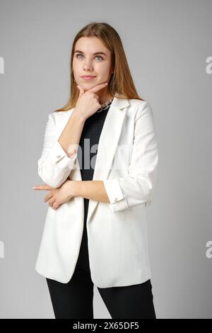 Thoughtful Young Woman in a Stylish White Blazer Posing on gray background Stock Photo