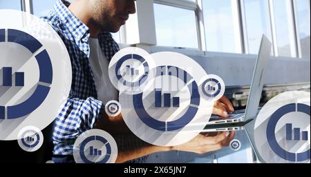 Image of graph icon in circles over latino man typing on keyboard of laptop in office Stock Photo