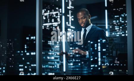 Portrait of Stylish Black Businessman Using Laptop, Looking out of Window on a City. successful African-American CEO Working on Computer, Working Hard Late At Night to Achieve Best Results Stock Photo