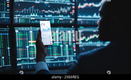 Financial Analyst Using Smartphone with Real-Time Stocks, Commodities and Market Charts. Working on Multi-Monitor Workstation with Stock Charts. Businessman Works in Investment Bank in the Evening. Stock Photo