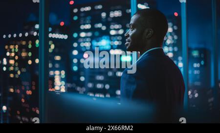 Successful Businessman Looking Out of the Window on Late Evening. Modern Hedge Fund Investor Enjoying Successful Life. Urban View with Down Town Street with Skyscrapers at Night with Neon Lights. Stock Photo