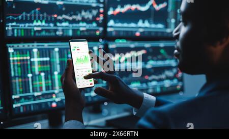 Financial Analyst Using Smartphone with Real-Time Stocks, Commodities and Market Charts. Working on Multi-Monitor Workstation with Stock Charts. Businessman Works in Investment Bank in the Evening. Stock Photo