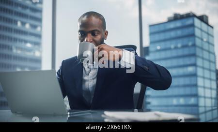 Portrait of Black Businessman in Tailored Suit Working on Laptop Computer and Drinking Coffee in His City Office. Digital Entrepreneur does Analysis, Cloud Computing for e-Commerce Strategy assessment Stock Photo