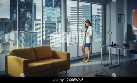 Successful Caucasian Businesswoman Using Smartphone While Standing in Office Looking out of Window on Big City. Confident Female Digital Entrepreneur Developing e-Commerce Software Strategy. Stock Photo