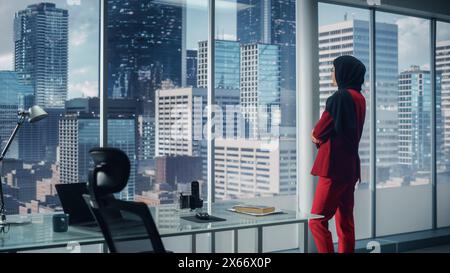 Successful Muslim Businesswoman Wearing Suit and Burka Standing in Office Looking out of Window on Big City. Confident Female Digital Entrepreneur Planning Investment Strategy for e-Commerce Startup Stock Photo