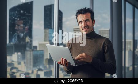 Portrait of Charismatic Businessman Using Laptop, Looking at Camera while Standing in Big City Office. Successful Software Engineer doing Data Analysis for His e-Commerce Startup Investment Stock Photo