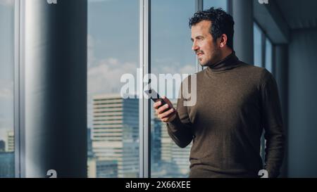 Thoughtful Arab Businessman Using Smartphone Standing in Office Looking out of the Window on Big City. Charismatic Digital Entrepreneur Plan Investment Strategy for e-Commerce Startup. Stock Photo