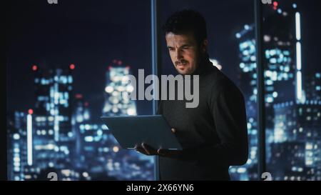 Big City Modern Office at Night: Successful Businessman Standing and Using Laptop. Handsome Male Digital Entrepreneur Thinking of Investment Strategy for e-Commerce Project. Stock Photo