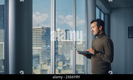 Thoughtful Middle Eastern Businessman Using Laptop while Standing in Office Near Window on Big City. Successful Digital Entrepreneur Doing Data Analysis management for e-Commerce Investment Stock Photo