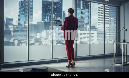 Successful Caucasian Businesswoman Using Laptop While Standing in Office Looking out of Window on Big City. Confident Female Managing Environmental, Social and Corporate Governance Stock Photo