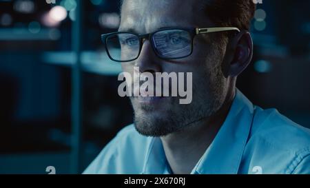 At Night Portrait of Handsome Startup Digital Entrepreneur Working on Computer, Line of Code Reflecting in Glasses. Developer Working on Innovative e-Commerce App using AI Algorithm Big Data. Blue Stock Photo