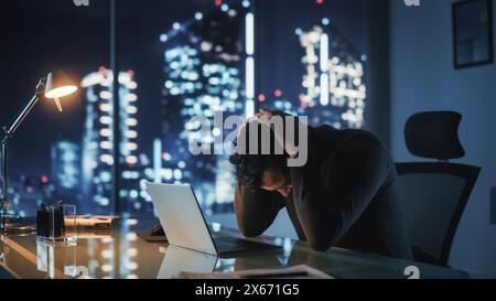 Portrait of Stressed Businessman Working on Laptop Computer in His Big City Office at Night. Overworked Tired Entrepreneur trying to Find Solution for Business Problems. Stock Photo