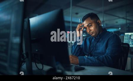 Night Office: Portrait of Handsome Indian Man in Working on Desktop Computer. Digital Entrepreneur Typing, Creating Software, e-Commerce App Design, Programming. Thoughtful Man Seeking Solution Stock Photo