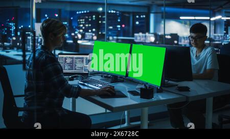 Diverse Teamwork in Office at Night: Person with Disability Using Prosthetic Arm to Work on Green Screen Chroma Key Computer. Team of Software Engineers work Late to create Innovative App Stock Photo