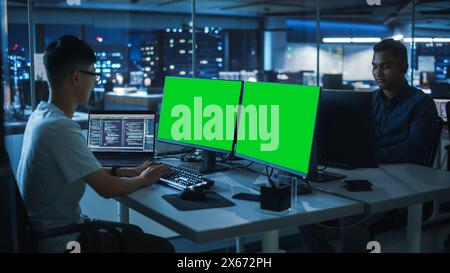 Night Office: Young Japanese Man in Working on Green Screen Chroma Key Desktop Computer. Team of Programmers Typing Code, Creating Modern Software, e-Commerce App Design, e-Business Programming Stock Photo