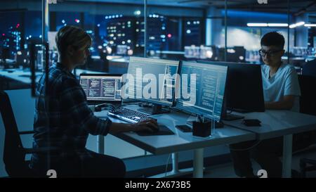 Diverse Teamwork in Office at Night: Motivated Woman with Disability Using Prosthetic Arm to Work on Computer. Specialist uses Myoelectric Bionic Hand. Software Engineers work Late on Innovative App Stock Photo
