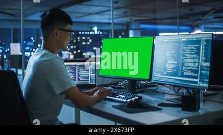 Night Office: Young Japanese Man in Working on Green Screen Chroma Key Desktop Computer. Programmer Typing Code, Creating Modern Software, e-Commerce App Design. Over Shoulder Stock Photo