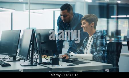 Teamwork In Diverse Inclusive Office: Project Manager Talks with Woman with Disability with Prosthetic Arm to Work on Desktop Computer. Professionals Software Engineers Create e-Commerce App Solution Stock Photo