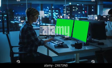 Diverse Teamwork in Office at Night: Person with Disability Using Prosthetic Arm to Work on Green Screen Chroma Key Computer. Team of Software Engineers work Late to create Innovative App Stock Photo