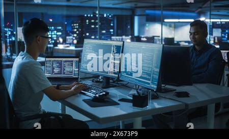 Night Office: Young Japanese Man in Working on Desktop Computer. Diverse Multi-Ethnic Team of Programmers Typing Code, Creating Modern Software, e-Commerce App Design, e-Business Programming. Stock Photo