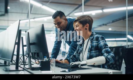 Teamwork In Diverse Office: Smiling Project Manager Talks with Happy Woman with Disability with Prosthetic Arm to Work on Desktop Computer. Professionals Create e-Commerce Software App Solution Stock Photo