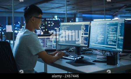 Night Office: Young Japanese Man in Working on Desktop Computer. Digital Entrepreneur Typing Code, Creating Modern Software, e-Commerce App Design, e-Business Programming. Over Shoulder Stock Photo