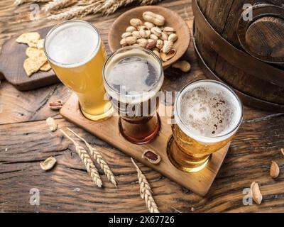 Three craft beers on wooden beer flight board. Top view of beer glasses, snacks and beer cask at the background. Stock Photo
