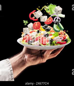 Cook holds white plate of greek salad on black background. Ingredients of salad falling down om the plate. Tasty food background. Stock Photo