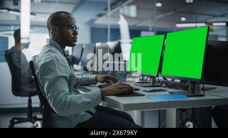 Multi-Ethnic Office: Black IT Programmer Working on Computer with Green Screen Chroma Key Display. Male Software Engineer Developing App, Program, Video Game. Terminal with Code Language. Stock Photo