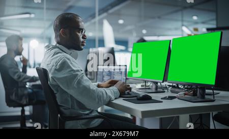 Multi-Ethnic Office: Black IT Programmer Working on Computer with Green Screen Chroma Key Display. Male Software Engineer Developing App, Program, Video Game. Terminal with Code Language. Stock Photo