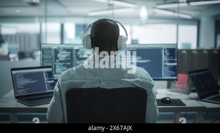 Office: Professional Black IT Programmer Uses Headphones while Working on Desktop Computer. Male Website Developer, Software Engineer Developing App, Video Game. Listening to Podcast, Music. Back View Stock Photo