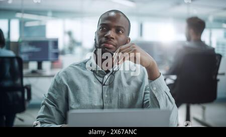 Modern Office: Portrait of Thoughtful Black IT Programmer Working on Laptop, Finding Solution, Solving Problems, Brainstorming. Male Software Engineer Wearing Glasses Develop App, Program, Video Game Stock Photo