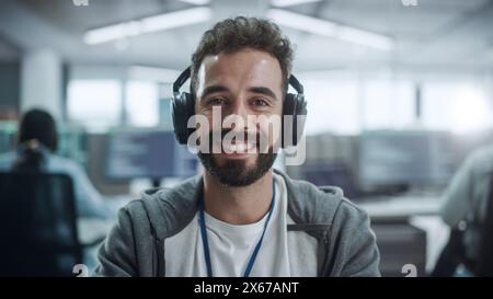 Office: Portrait of Happy IT Programmer Wearing Headphones Working on Desktop Computer, Looking at Camera and Smiling. Male Software Engineer Developing App, Video Game. Listening to Podcast, Music Stock Photo