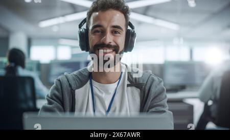 Office: Portrait of Happy IT Programmer Wearing Headphones Working on Desktop Computer, Looking at Camera and Smiling. Male Software Engineer Developing App, Video Game. Listening to Podcast, Music Stock Photo