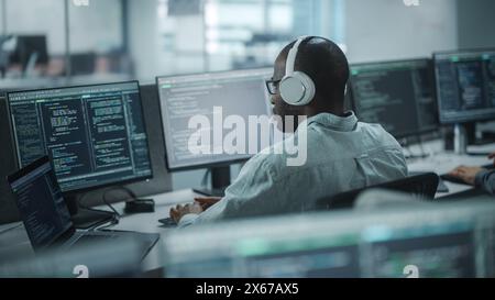 Office: Professional Black IT Programmer Uses Headphones while Working on Desktop Computer. Male Website Developer, Software Engineer Developing App, Video Game. Listening to Podcast, Music. Stock Photo