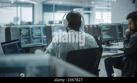 Office: Professional Black IT Programmer Uses Headphones while Working on Desktop Computer. Male Website Developer, Software Engineer Developing App, Video Game. Listening to Podcast, Music. Stock Photo
