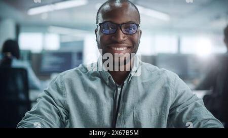 Office: Portrait of Focused IT Programmer Wearing Headphones Working on Computer, Looking at Camera and Smiling. Black Male Software Engineer Developing Innovative App, Program, Video Game. Stock Photo