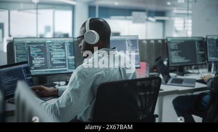 Office: Professional Black IT Programmer Uses Headphones while Working on Desktop Computer. Male Website Developer, Software Engineer Developing App, Video Game. Listening to Podcast, Music. Stock Photo