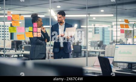 Young Businessman Talking with Experienced Colleague while Using Laptop Computer in Office. Employees Discuss Corporate, Financial and Marketing Projects. Specialists Work in Diverse Team. Stock Photo