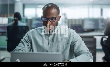 Modern Office: Portrait of Thoughtful Black IT Programmer Working on Laptop, Finding Solution, Solving Problems, Brainstorming. Male Software Engineer Wearing Glasses Develop App, Program, Video Game Stock Photo